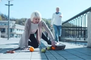 An elderly woman suffers a slip, trip, and fall injury and spills her shopping on the street while a bystander runs to help.