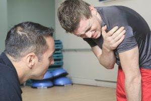 A man in a gym with his trainer, holding his shoulder in pain.