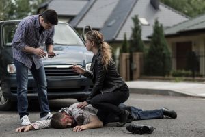 A car accident victim lies injured on the road. One person kneels next to them and another person is stood on the other side and holding a mobile phone.