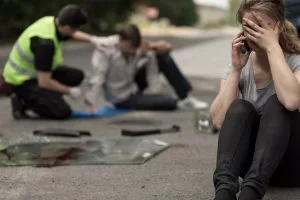 A person sat down on a road with a phone to their ear and one hand over their face. In the background, a person in a hi-viz jacket comforts a person who was injured in a car accident.