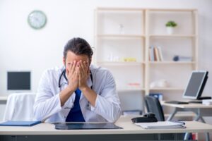 Male Doctor Sat At A Desk With Hands Covering His Face After Giving Negligent Treatment. 