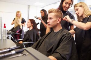 A Hairdresser Cuts A Man's Hair While Other Clients Have Their Hair Dried And Brushed In the Background. 