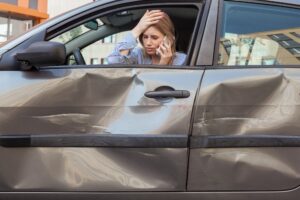 A white woman talks on the phone and holds her head in distress. The side of her car is damaged.