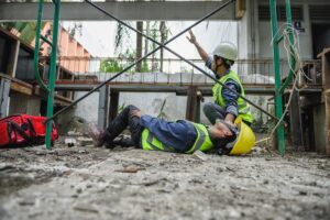 A construction worker in a high vis vest lays on the ground as a coworker signals for first aid