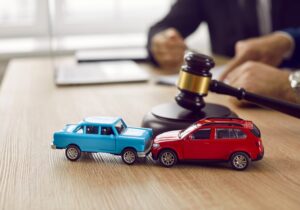 Two Toy Cars Crashed Into Each Other In Front Of Gavel With Out Of Focus Lawyer At Wooden Desk. 