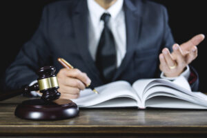 A lawyer in a suit sat at a desk, shot from the shoulders down. The solicitor is writing in an open book and there is a gavel in front of the book.