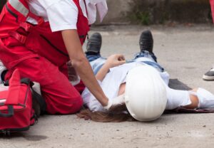 2 workers in red overalls. One is female ang lying down on her back, and the other is crouched beside her with their hand on her left shoulder. 