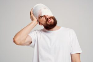 A man in a white t-shirt, tilting his head to the side and holding it in pain. There are white bandages around his head and left eye. 