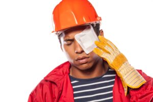 Male construction worker wearing yellow gloves and an orange helmet, holding his left eye in pain that has a white square plaster on.
