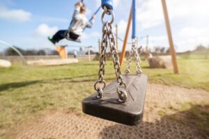 An empty swing set in the foreground with blurred grass and children playing in the background