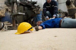 A man in blue overalls lying on the ground in a workshop with his colleague performing first aid